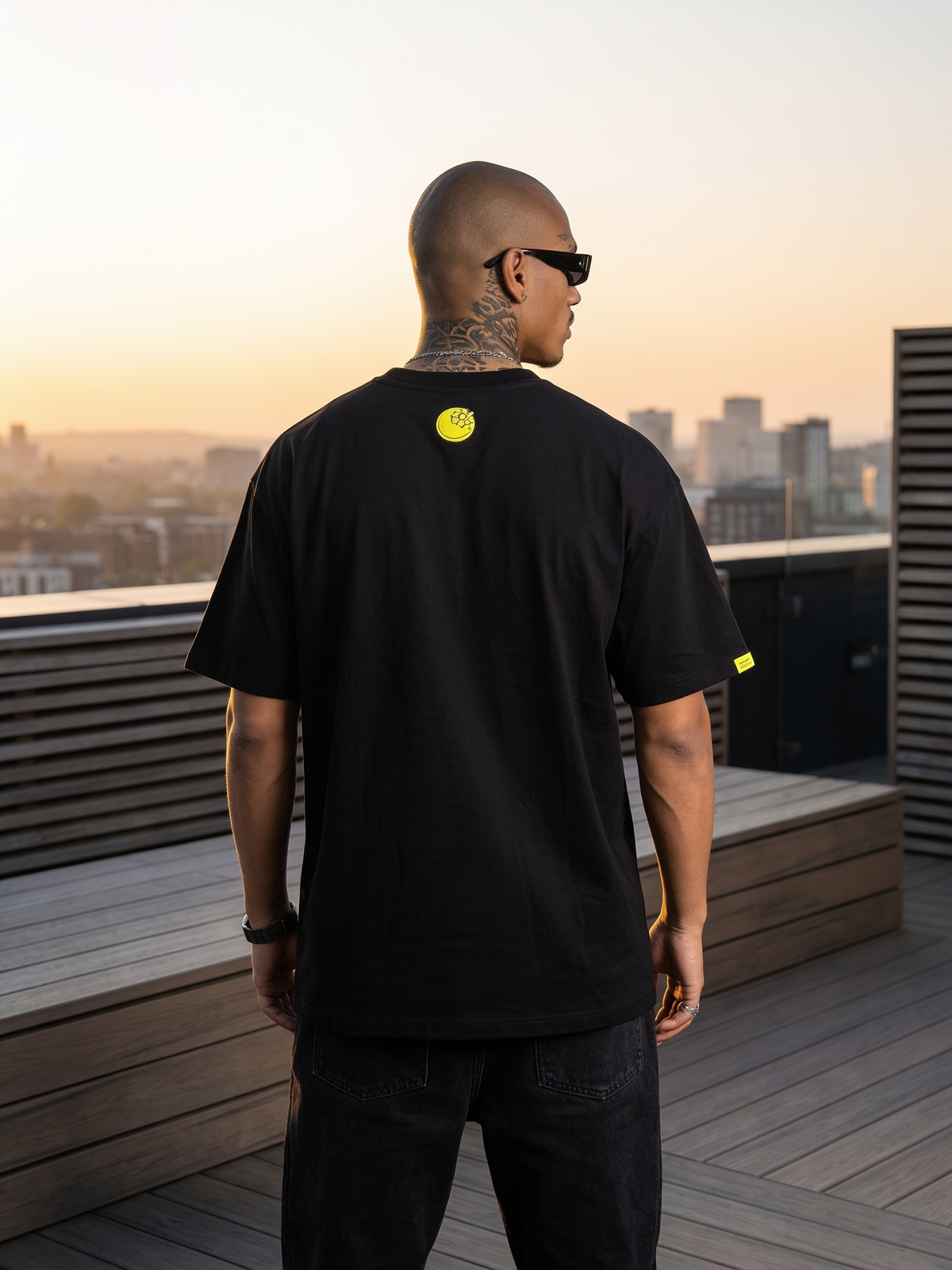 A man wearing a black t-shirt with a small yellow smiley face logo on the back, standing on a wooden deck at sunset with a city skyline in the background.
