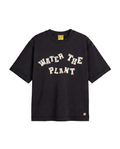 A black short-sleeve t-shirt with 'WATER THE PLANT' charms across the chest, featuring a small yellow flower logo on the bottom right hem and a yellow brand label inside the collar.