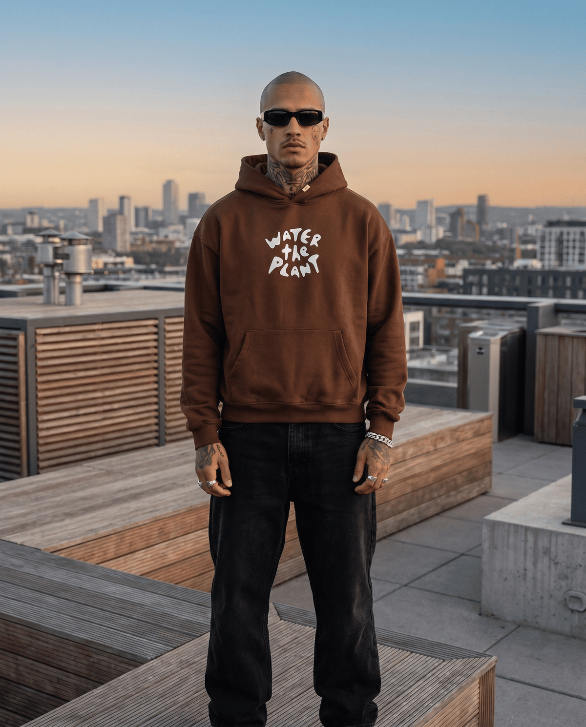 Man wearing a brown hoodie with 'Water the Plant' text on a rooftop with cityscape background
