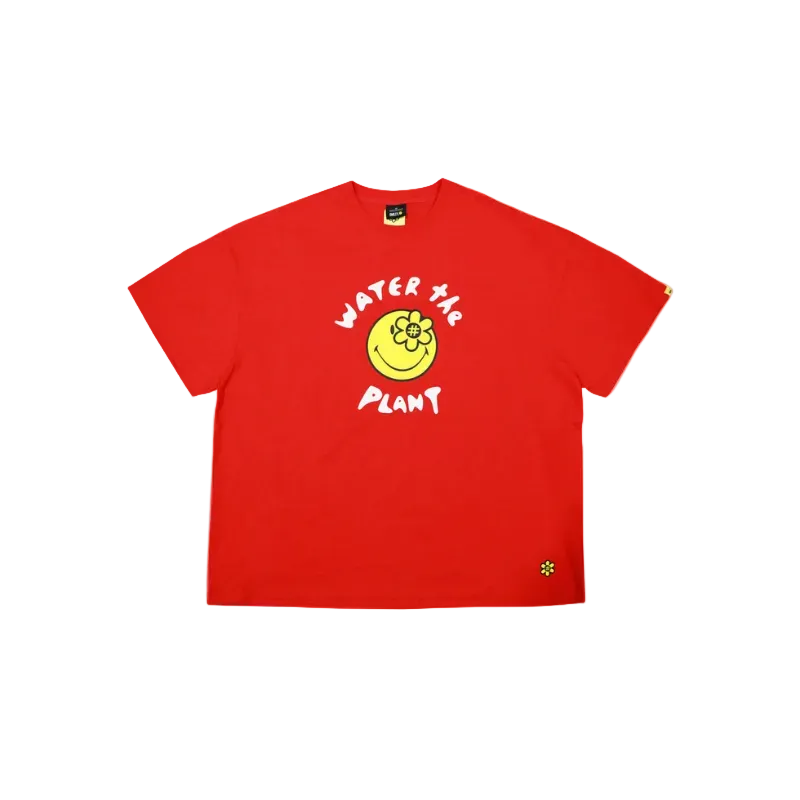 A bright red short-sleeve t-shirt with a graphic on the chest featuring a yellow smiley face with a flower and the words 'WATER THE PLANT' in white. A small yellow flower logo is printed on the bottom right hem.