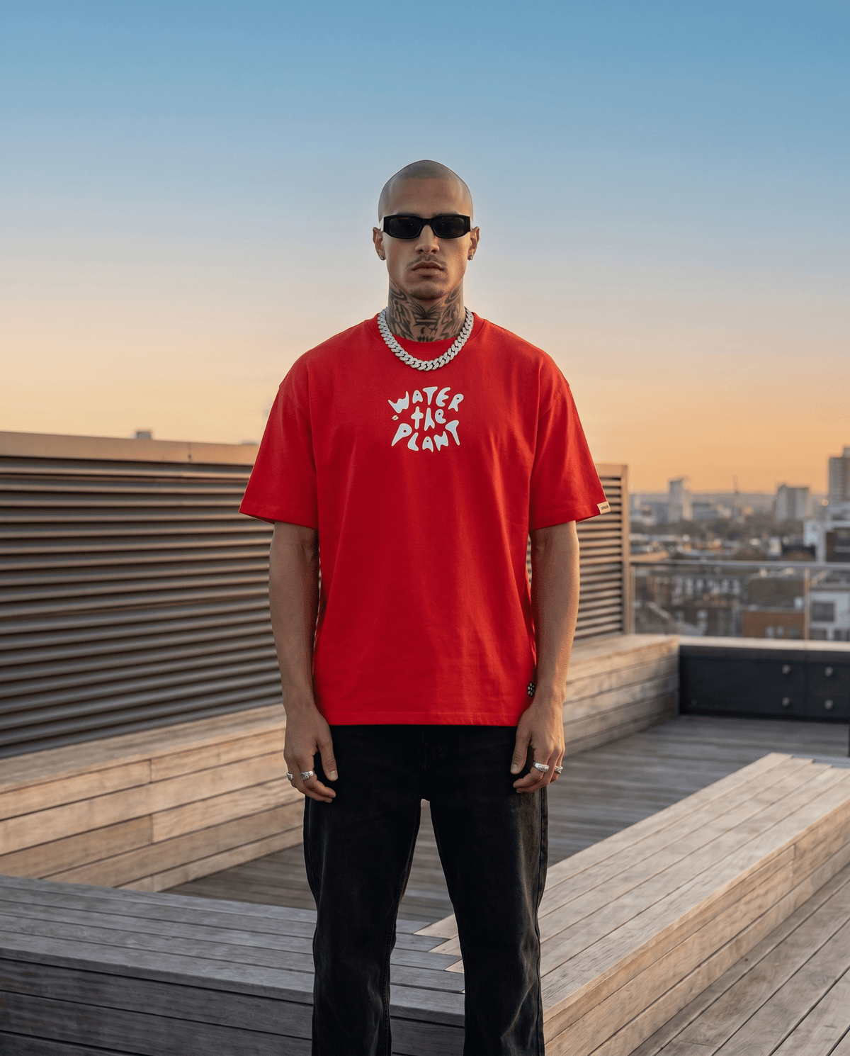 A man wearing a bright red crewneck t-shirt with a white graphic print that reads 'WATER THE PLANT' stands on a wooden rooftop terrace at sunset.