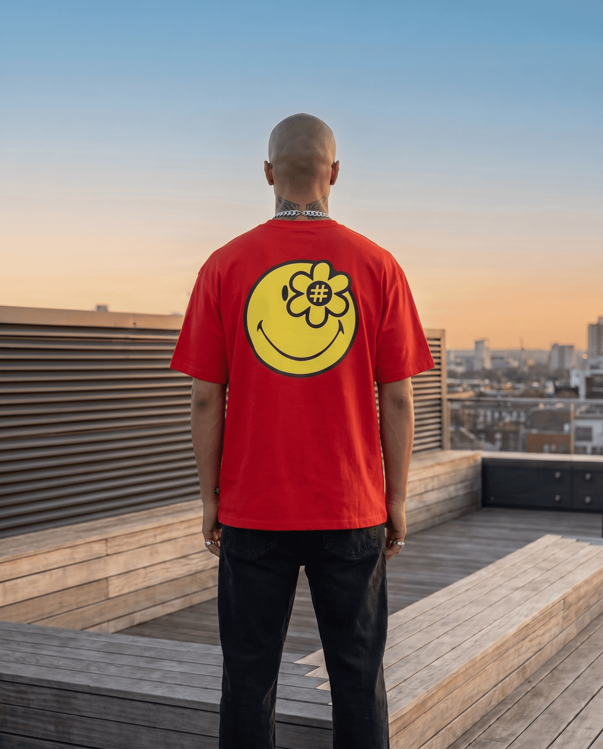 A man viewed from behind wearing a bright red t-shirt with a large yellow smiley® face graphic on the back, standing on a wooden rooftop terrace at sunset.