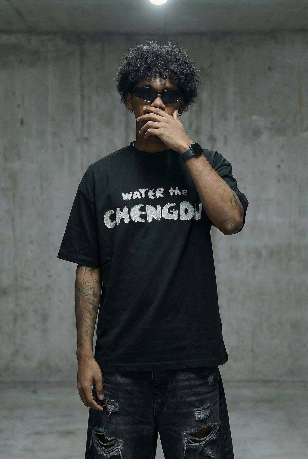 A man wearing a black graphic t-shirt with the text 'WATER the CHENGDU' in white, paired with ripped black jeans and sunglasses, standing against a concrete wall.