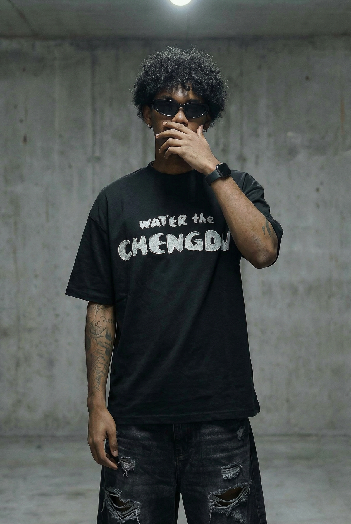 A man wearing a black graphic t-shirt with the text 'WATER the CHENGDU' in white, paired with ripped black jeans and sunglasses, standing against a concrete wall.