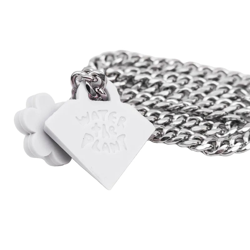 A silver-toned metal chain necklace with a small square pendant engraved with 'WATER THE PLANT', shown next to a white flower-shaped charm on a plain white background.