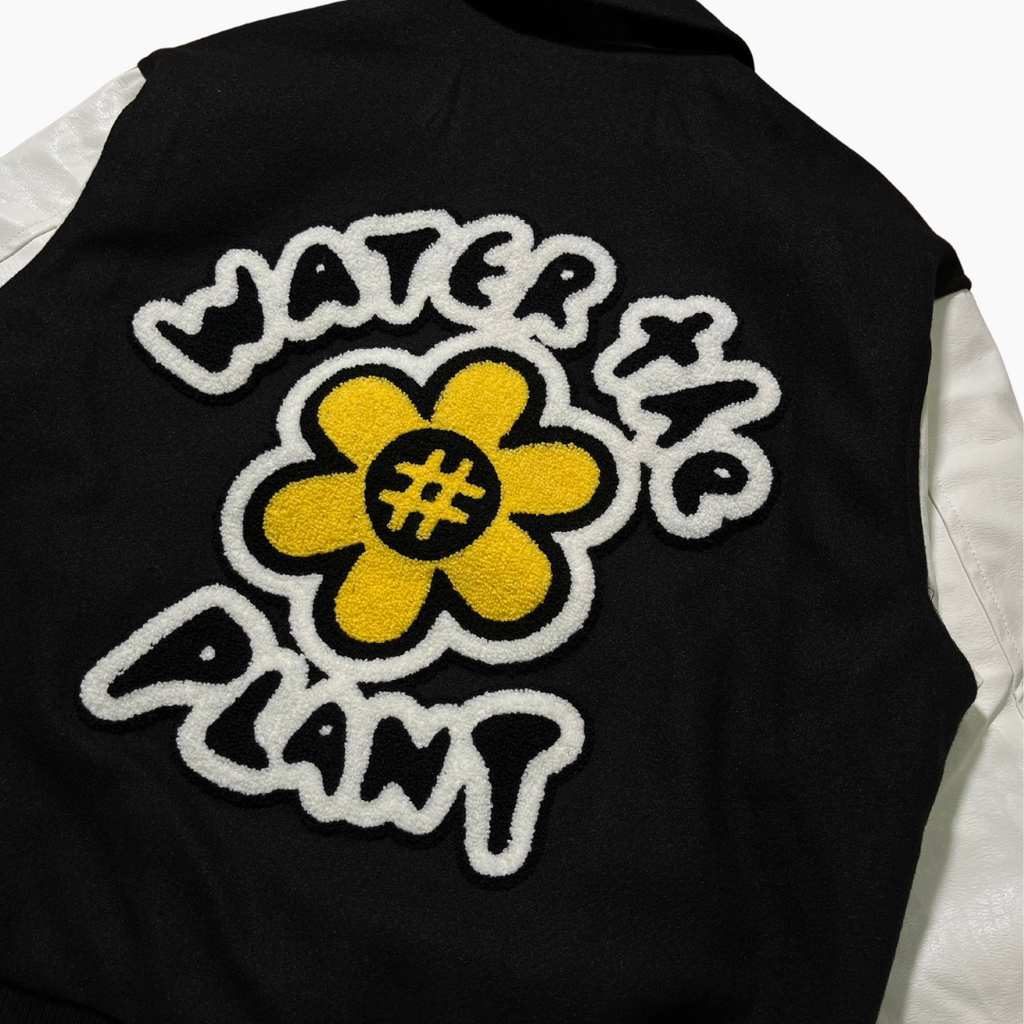 Close-up of the back of a black varsity-style jacket with white sleeves, featuring a large raised yellow flower patch with a black hashtag in the center and white fuzzy text reading 'WATER THE PLANT' around it.