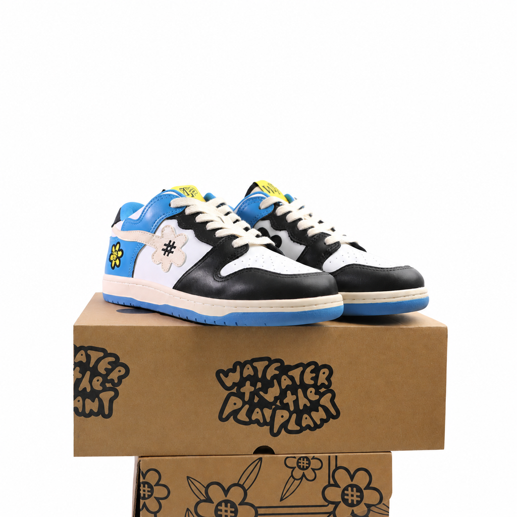 A pair of low-top sneakers in black, white, and blue, displayed on top of a brown cardboard shoebox with floral graphics. The shoes feature a white side panel with a black hashtag symbol, a yellow flower patch, and a blue midsole.